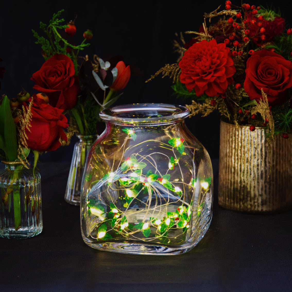 An image of a dark table with vases with red floral arrangements on them with a glass jar with christmas holly led string lights inside.