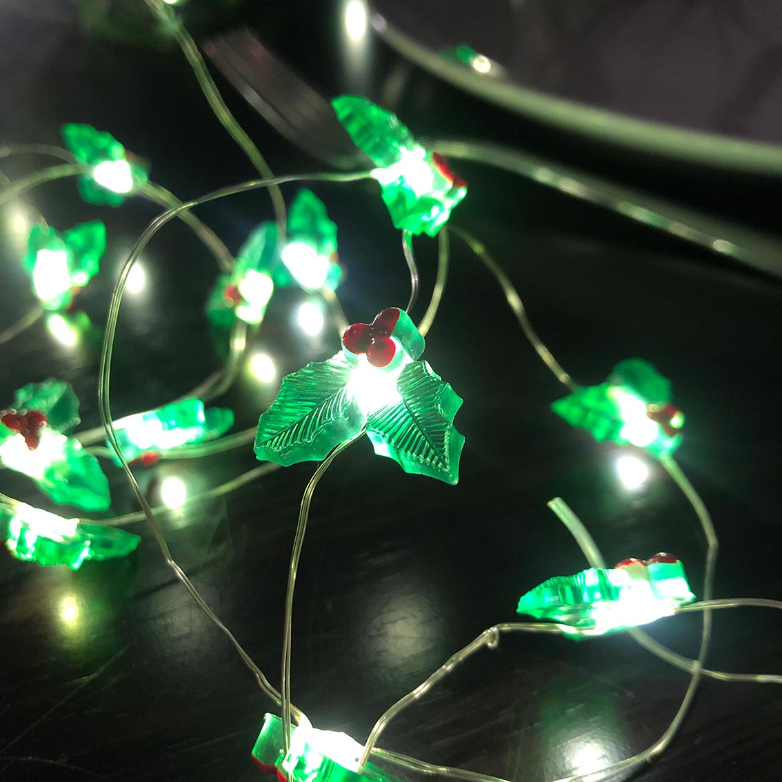 An image of christmas holly LED string lights glowing against a dark surface. They are green holly leaf shape with 3 red berries.