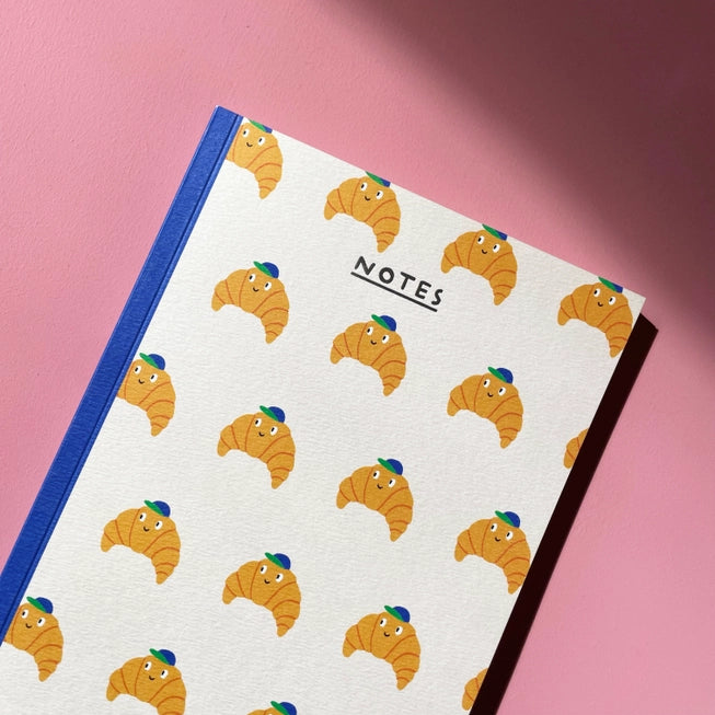 An image of a cream coloured notebook covered in illustrations of croissants with a baseball cap on and face. The notebook has a navy binding and the word &#39;notes&#39; written in silver foil.